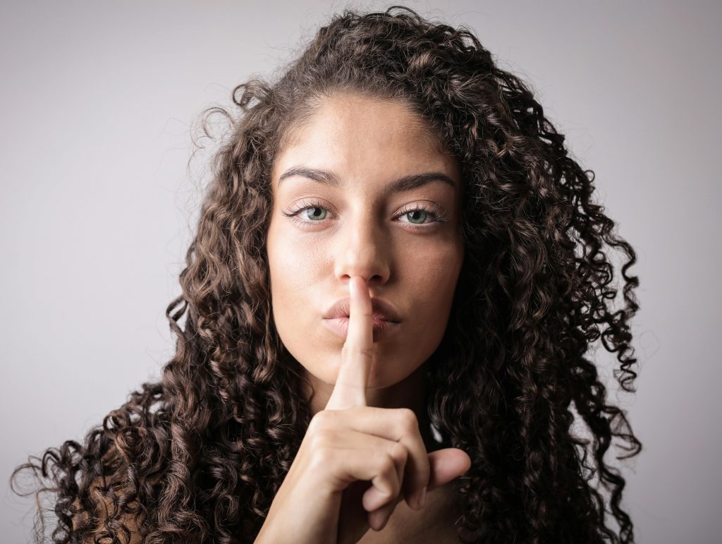 Close-up portrait of a woman with curly hair, signaling quiet with a finger on lips.